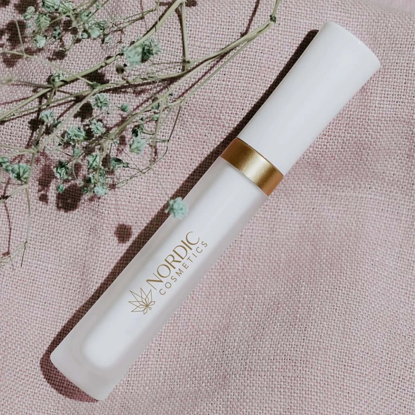 das Wimpernserum liegt auf einem pinken Tuch während ein kleiner Blumenstrauch dadrüber liegt