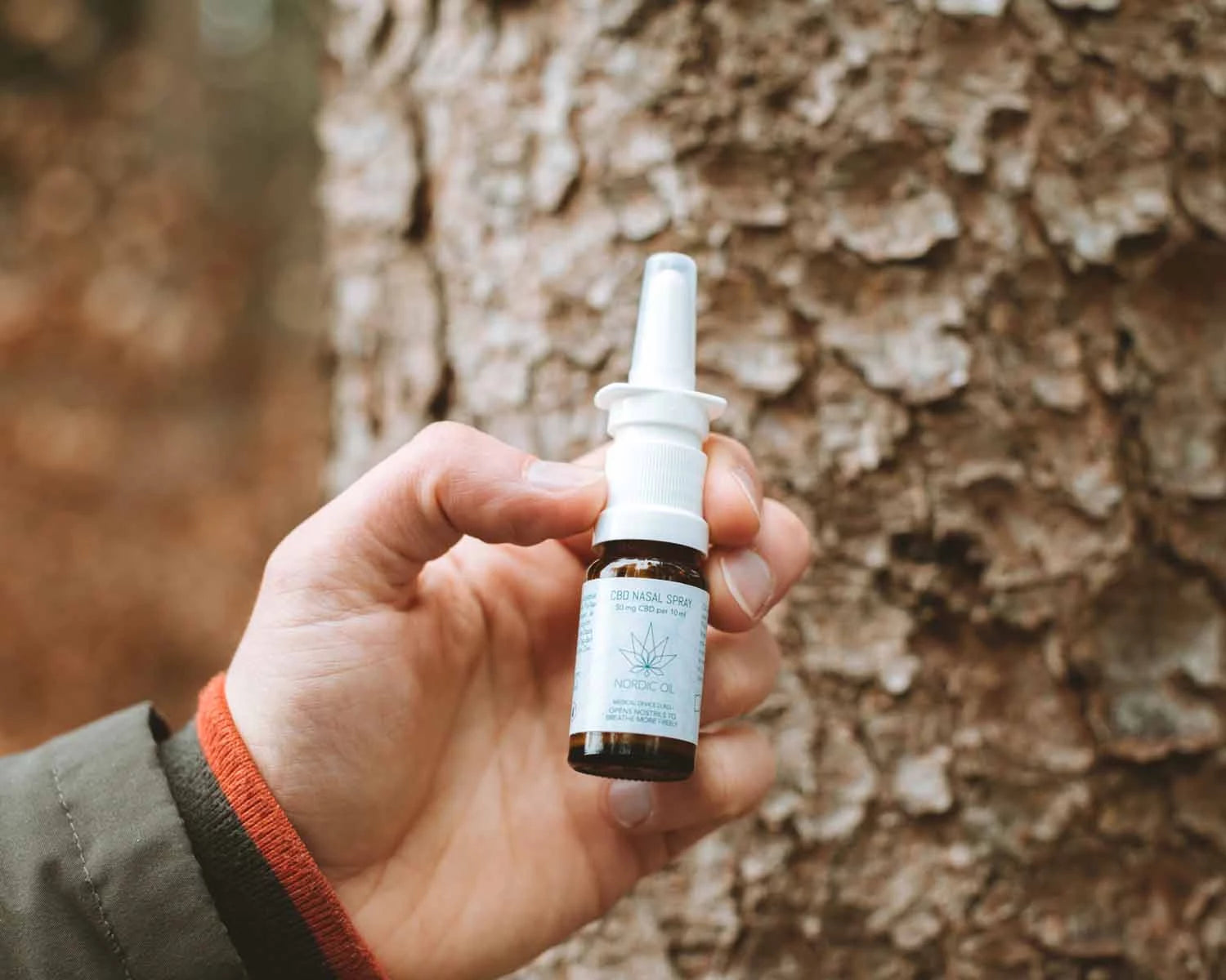 Eine Hand hält das Nordic Oil CBD Nasenspray vor einen Baum
