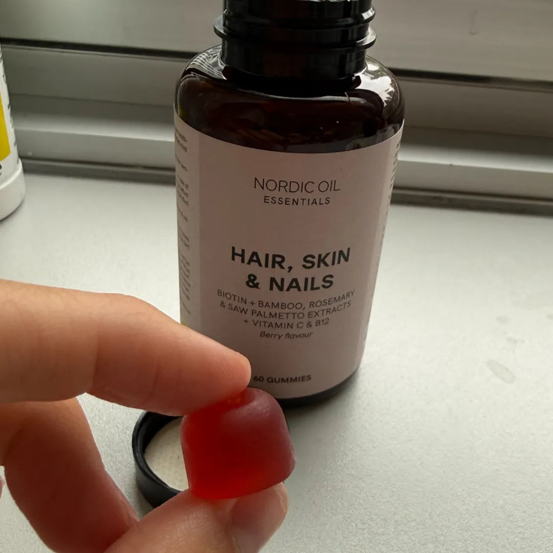 Rotes Gummibärchen wird zwischen Fingern gehalten, vor einer Nordic Oil Essentials Hair, Skin & Nails Gummies-Flasche.