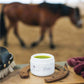 CBD Balsam steht in einem Hufeisen neben einer Bürste für Pferde, während ein braunes Pferd im Hintergrund auf einer Koppel trabt